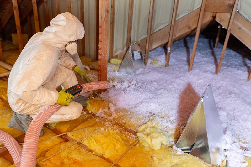 A worker in a protective suit and respirator uses a hose to blow loose-fill insulation into an attic.